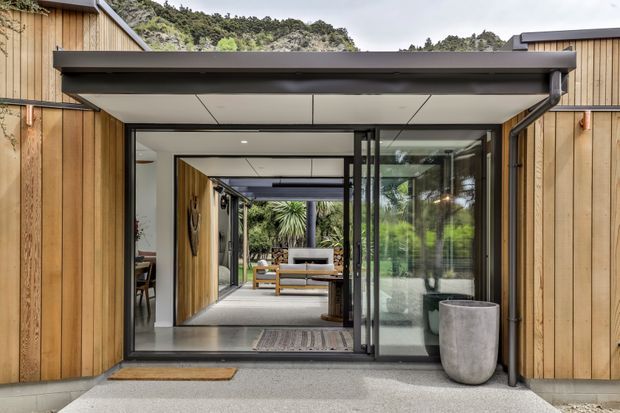 A contemporary timber-clad house entrance with a deep flat-roofed canopy and fully glazed sliding doors. The view looks through the entry into a bright open-plan interior, with exposed dark framing, concrete paving, warm vertical timber wall cladding, and lush planting beyond. A small seating area and layered circulation spaces are visible inside.