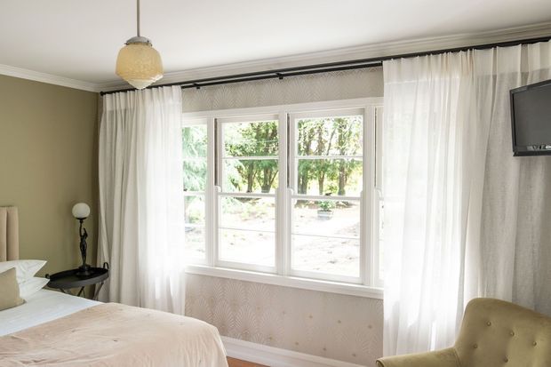 A softly styled bedroom with a large white-framed multi-pane window, sheer white curtains layered over patterned drapes, a pale upholstered bed, and a tufted armchair. The room has muted taupe walls, light bedding, a wall-mounted television, and a warm pendant light, creating a calm, elegant atmosphere.