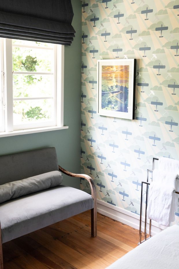 A compact, thoughtfully styled bedroom or sitting area with a grey upholstered daybed, curved timber arm, dark Roman blind, and warm timber flooring. One wall is covered in playful pale wallpaper featuring repeated blue-grey aircraft motifs, while the adjacent wall is painted muted sage green. A framed artwork, white-trimmed window, and freestanding towel or textile rack complete the room.
