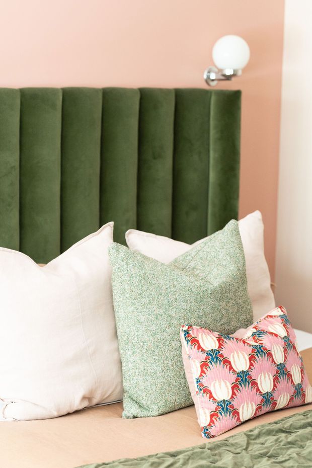 Close-up view of a styled bedroom featuring a tall vertically channelled deep green upholstered headboard, soft blush-pink walls, pale bedding, mint-green textured pillows, and a colourful floral accent cushion. A small spherical wall light with chrome detailing is mounted above the headboard. The room has a playful, colourful, softly layered interior aesthetic.