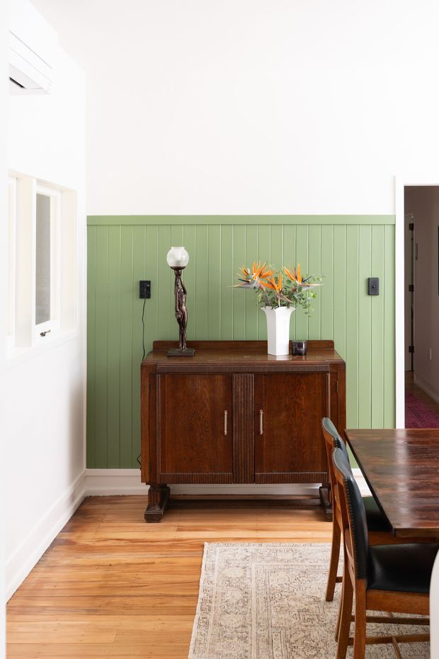 Bright dining area with white walls and a green vertical-panel wainscoted feature wall. A dark wood vintage sideboard is styled with a lamp and floral arrangement, while a matching dark wood dining table and chairs occupy the foreground. Timber flooring and a neutral patterned rug add warmth.