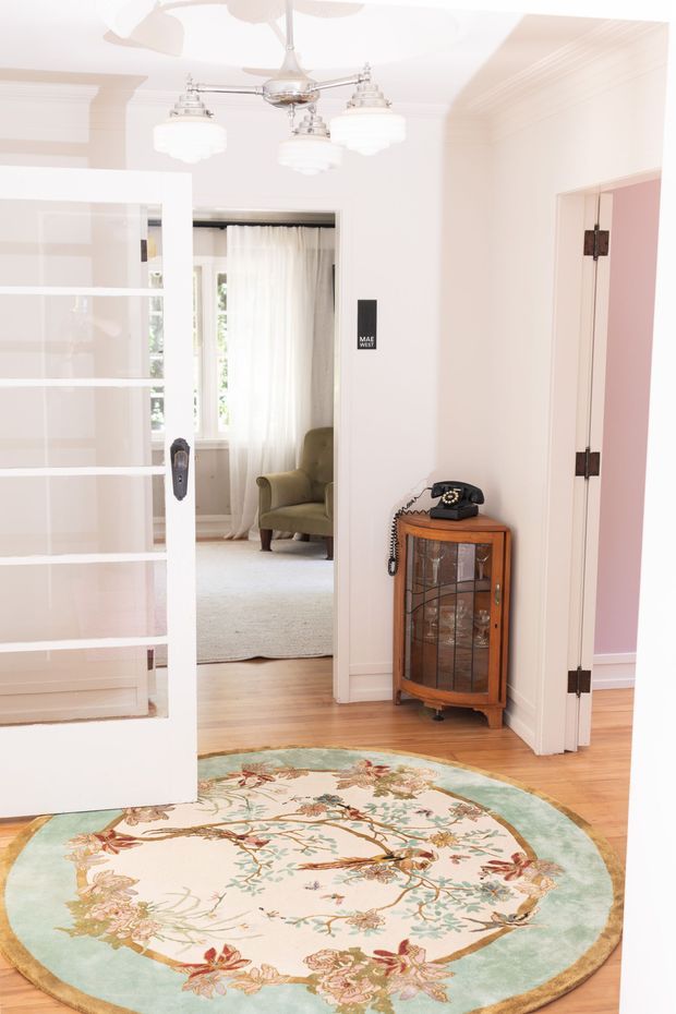 Bright, traditional-style residential hallway or entry area with white walls, decorative crown moulding, timber flooring, a vintage glass-front cabinet, rotary telephone, chandelier, and a floral round rug. A white glazed door opens toward a softly furnished adjoining room, while another doorway reveals a pink-toned room.