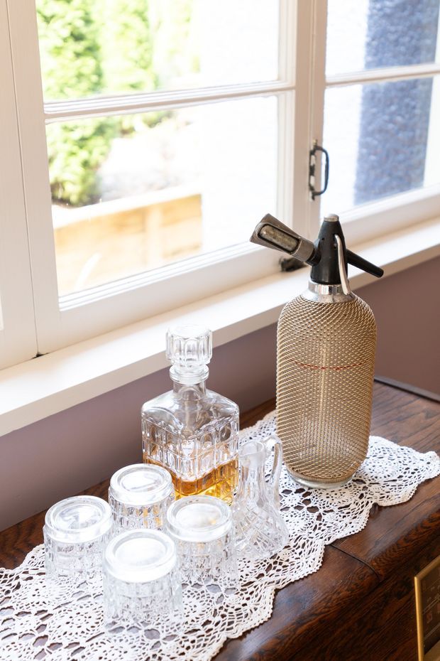 Close-up interior detail of a vintage-style drinks display on a dark timber sideboard beside a bright multi-pane window. The arrangement includes a textured metal soda siphon, crystal decanter and several cut-glass tumblers on a white crocheted lace runner. White-painted timber window frames and warm natural light create a nostalgic, traditional atmosphere.
