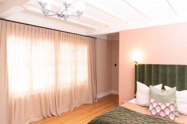 Bright, softly styled bedroom with blush-pink walls, warm timber flooring, full-height sheer curtains, and a tall channel-tufted olive green upholstered headboard. The bed is dressed in white bedding with decorative cushions and a textured dark green throw. A polished multi-arm ceiling light and wall sconce add a refined decorative touch.