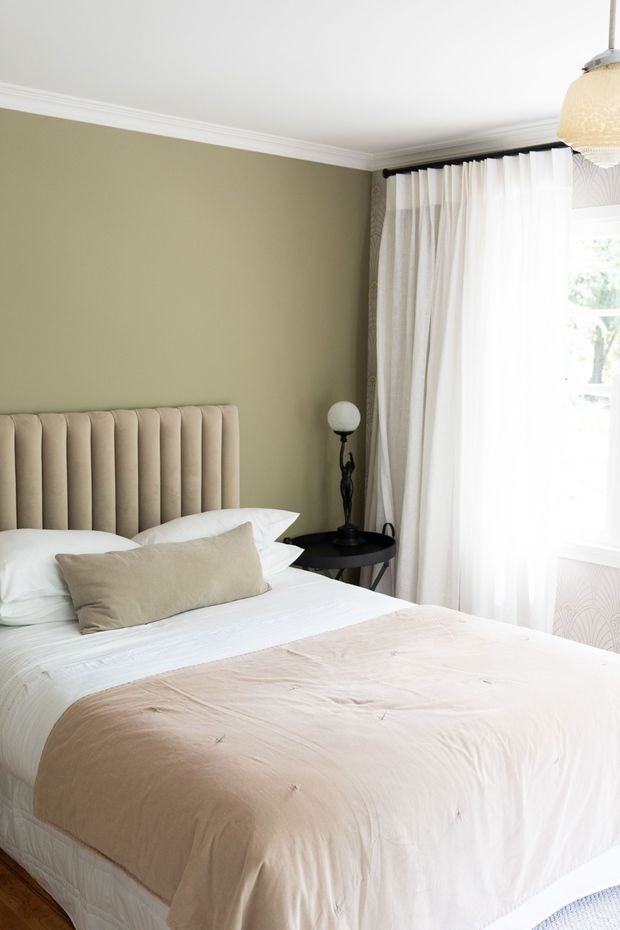 A softly styled bedroom with a large bed dressed in white and beige bedding, a tall vertically channelled upholstered headboard, and a muted sage-green wall. White crown moulding and sheer floor-length curtains frame a bright window. A small black round side table with a decorative lamp sits beside the bed, while a warm pendant light hangs near the upper right corner. The room has a calm, refined, neutral aesthetic.