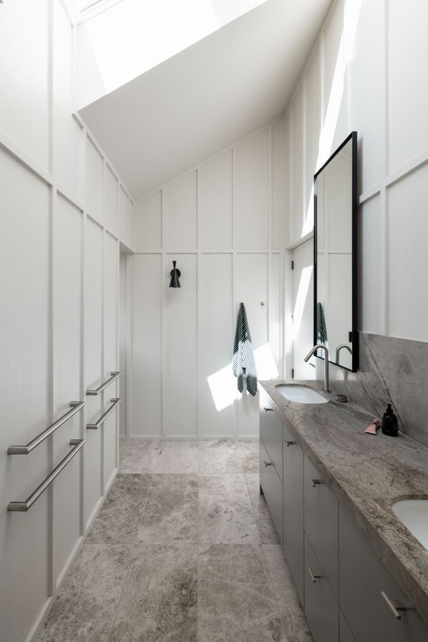 Bright, narrow contemporary bathroom with white vertical wall panelling, integrated cabinetry and long horizontal metal pulls on the left, a grey natural-stone vanity benchtop with multiple basins and brushed metal tapware on the right, large black-framed mirror, and pale stone-look floor tiles. Strong natural light enters from high windows, creating a clean, airy interior.