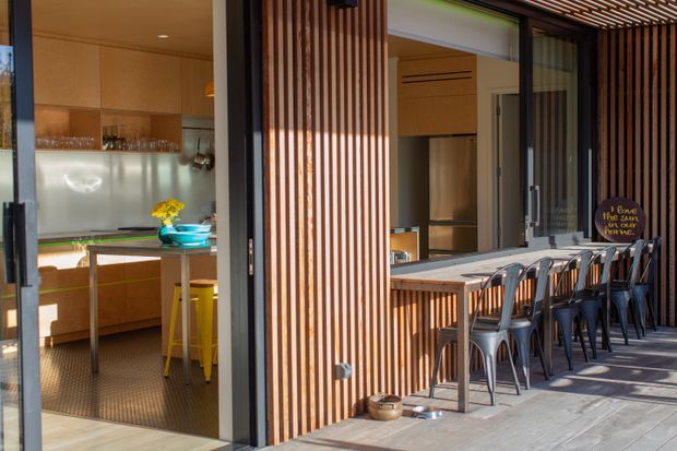 A contemporary indoor-outdoor entertaining area with a compact kitchen opening directly onto a covered bar and dining terrace. Warm timber cabinetry and vertical timber battens frame large glazed openings, with a long built-in counter and dark metal stools outside. The space is brightly lit and designed for casual entertaining.