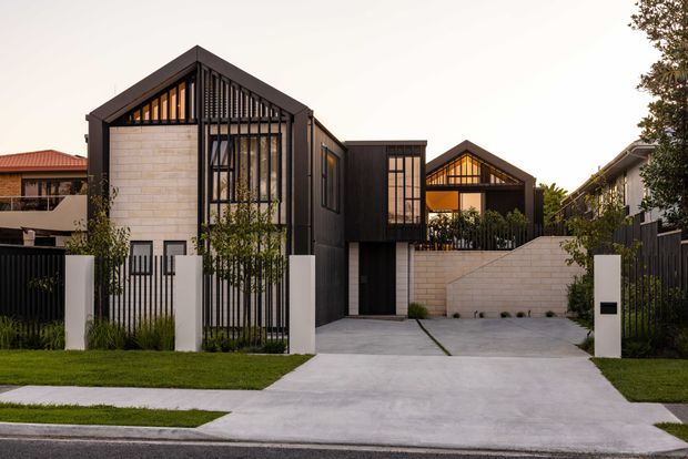 Street-facing view of a contemporary multi-level residential home with steep gabled roof forms, dark vertical cladding and metal screens, pale masonry walls, large glazed openings, and a broad concrete driveway. The landscaped frontage includes lawn, planting, and a dark perimeter fence.