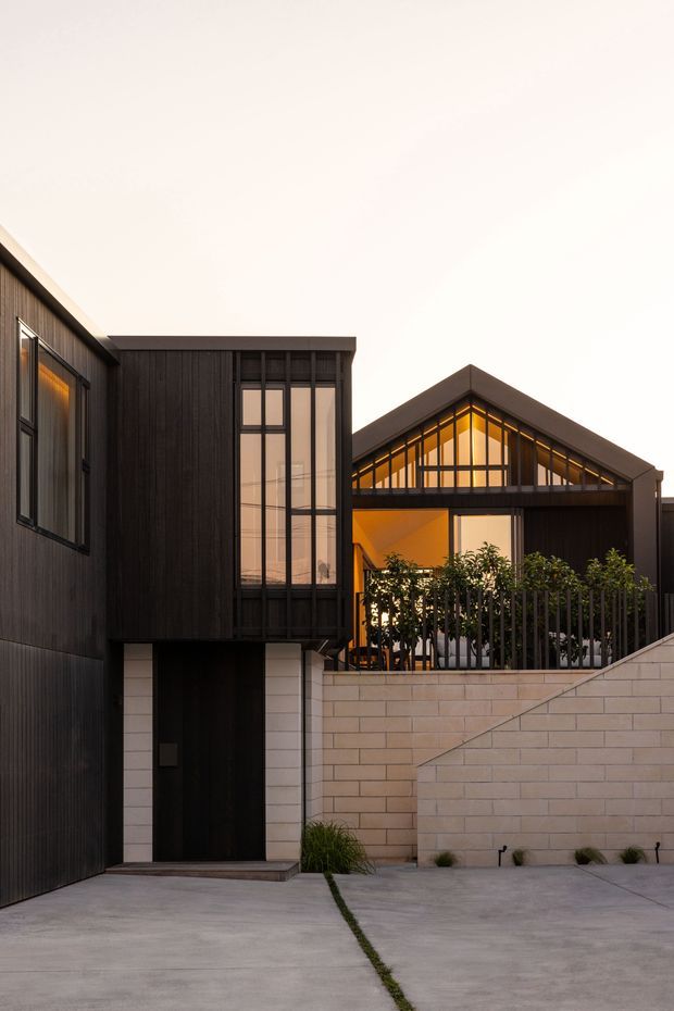 Contemporary residential facade viewed from the street, featuring dark vertical cladding, expansive black-framed glazing, a prominent gabled glazed section with warm interior lighting, a pale masonry boundary wall, and a concrete driveway or forecourt.