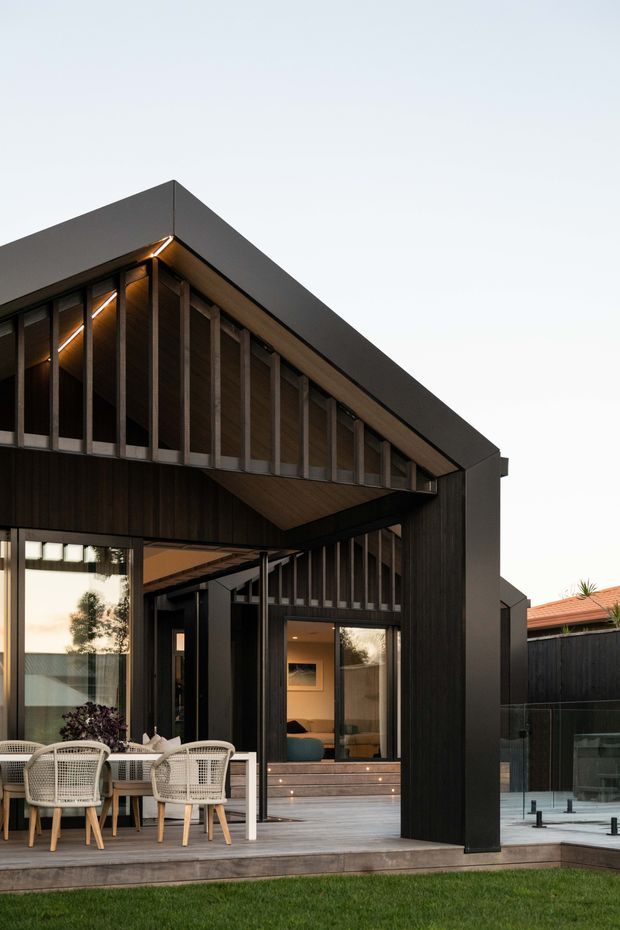 Contemporary dark-clad residential exterior with a strong gabled roof form, vertical timber-like screening, extensive glazing, and a covered outdoor entertaining area. A dining setting and interior living space are visible through the openings, with a paved pool or water feature area to the side.