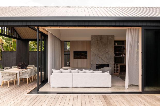 Contemporary open-plan living area opening onto a timber deck and covered outdoor dining space. The room features large retractable glazed doors, white sofas, warm timber wall panelling and ceiling finishes, a pale stone floor, and a dramatic vertically veined stone fireplace surround with integrated shelving and wine storage. Dark exterior framing provides contrast against the light, warm interior.