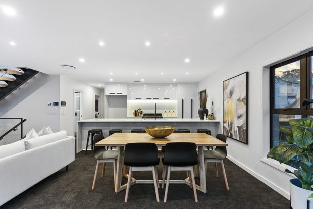 Bright open-plan interior combining a contemporary kitchen, dining area and living space. The room has white flat-panel cabinetry, a long island with a dark benchtop, a timber dining table with black upholstered chairs, a white sofa, recessed ceiling lights, dark carpet flooring, large artwork, and a black-framed window with indoor plants. A stairway is visible at the left.