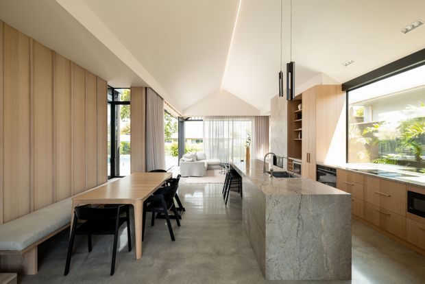 Bright open-plan kitchen and dining area with a long timber dining table and built-in bench seating, black dining chairs, timber cabinetry, a large stone island, integrated appliances, and pendant lighting. The space opens toward a glazed living area and garden, with polished concrete flooring and a sloped ceiling.