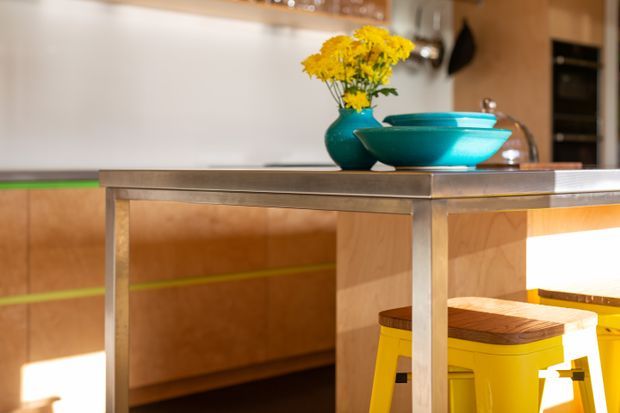 Close-up view of a contemporary kitchen or dining area featuring a stainless-steel-topped island or table with timber cabinetry below, bright yellow stools, and turquoise decorative bowls and vase holding yellow flowers. Warm wood finishes and vivid accent colours create a playful, colourful interior.