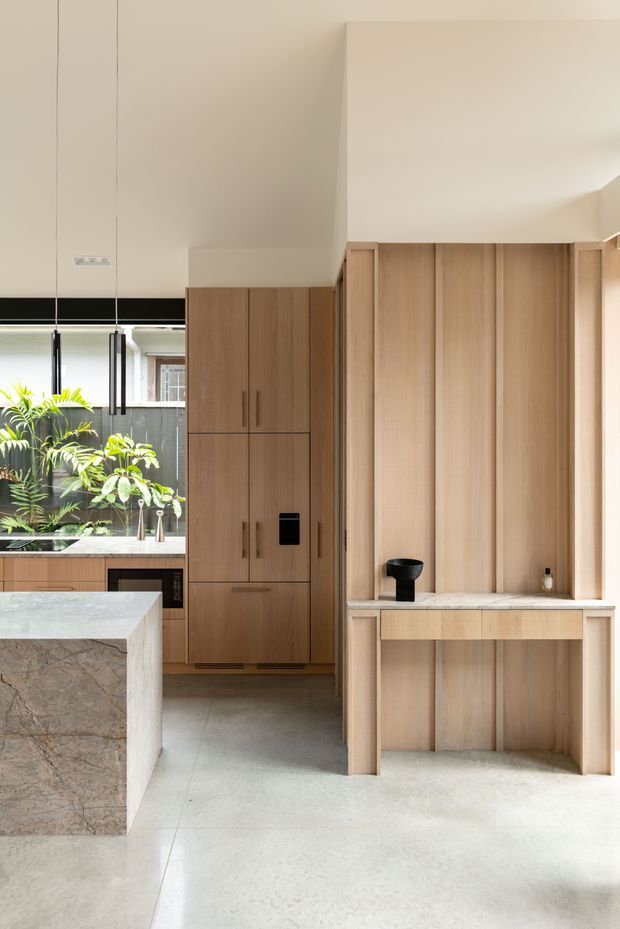 Bright contemporary kitchen interior with extensive light timber cabinetry, a large stone island, integrated appliances, and a built-in timber coffee or serving station. The space has a high ceiling, polished light concrete flooring, dark-framed glazing, and indoor planting visible near the rear.