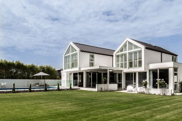 Exterior view of a large contemporary white two-storey residence with two prominent gabled wings, extensive floor-to-ceiling glazing and louvred window screens. A covered outdoor entertaining area opens onto a broad manicured lawn, with a swimming pool visible to the side.