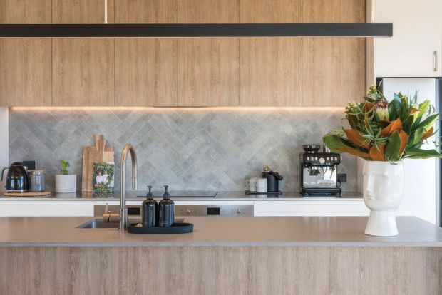 A contemporary kitchen viewed straight on, featuring a long island with an integrated sink and tall curved tap, light timber-look cabinetry, a pale stone-look benchtop, and a softly illuminated grey herringbone tile splashback. A black linear pendant spans the upper portion of the image, while coffee appliances and a large floral arrangement sit on the bench.