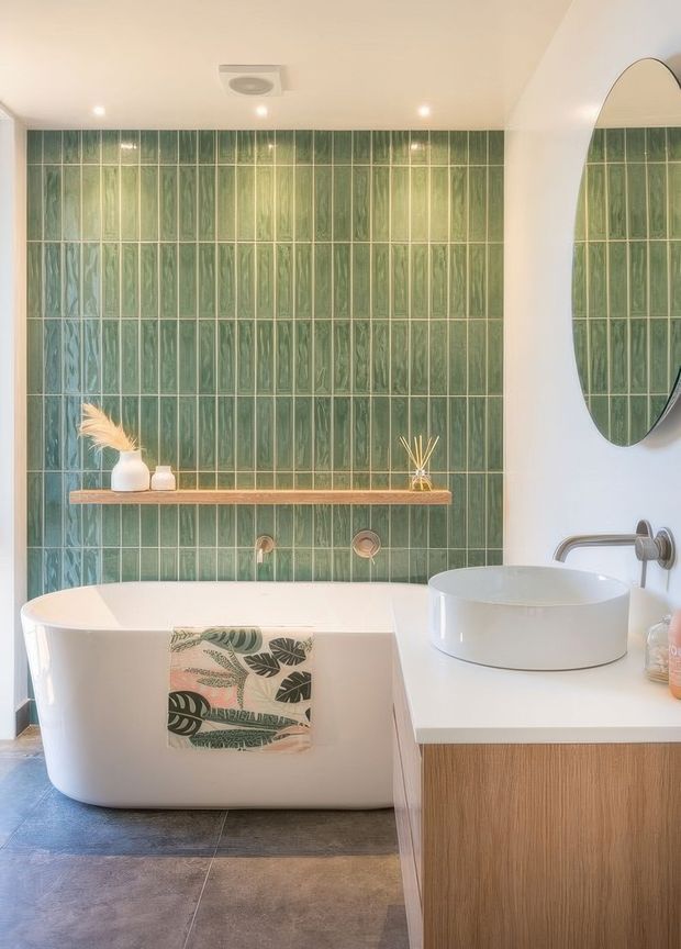 A bright, contemporary bathroom featuring a freestanding white bathtub, a round vessel basin on a timber-veneer vanity, and a wall-mounted curved tap. The rear wall is covered in vertically arranged glossy green textured tiles, complemented by a narrow timber shelf, an oval mirror, recessed ceiling spotlights, and dark stone-look floor tiles. Natural botanical accessories add a subtle organic accent.