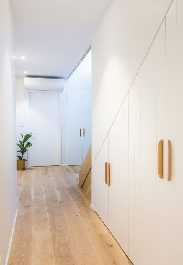 A bright, narrow residential hallway with pale timber flooring, white walls and ceiling, and extensive floor-to-ceiling built-in cabinetry along the right side. The cabinetry has simple vertical paneling and warm timber handles. A white entry door, additional storage cupboard, wall-mounted air conditioner, recessed lighting, and a small potted plant are visible at the end of the corridor.