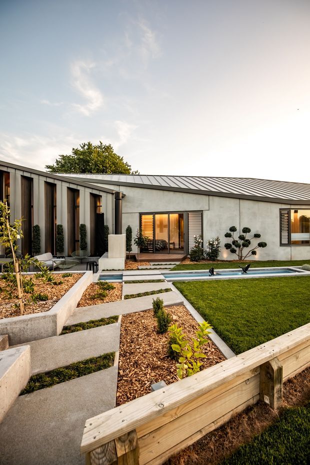 A landscaped courtyard garden viewed from outside a low, single-storey contemporary residence. The home has a long, shallow metal roof, pale rendered concrete walls, dark vertical window and door frames, and extensive glazing. Geometric concrete paths cross mulched planting beds and lead toward a narrow swimming pool. A manicured lawn, sculpted shrubs, raised timber garden edging, and warm interior lighting create a refined modern outdoor setting.