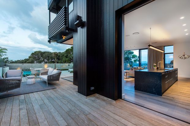 Contemporary residential exterior showing a spacious timber deck and outdoor seating beside a dark vertically clad house. Large open glazing connects the deck to a refined kitchen and living area, with a dark stone island, timber flooring, pendant lighting, and views toward a leafy landscape and water.