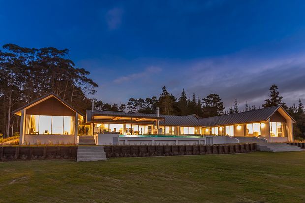 Twilight exterior view of a long, low-slung contemporary residence set in a landscaped lawn and surrounded by mature trees. The home features dark metal pitched roofs, extensive warm-lit glazing, timber-framed gabled end volumes, and a raised terrace with retaining walls and steps.