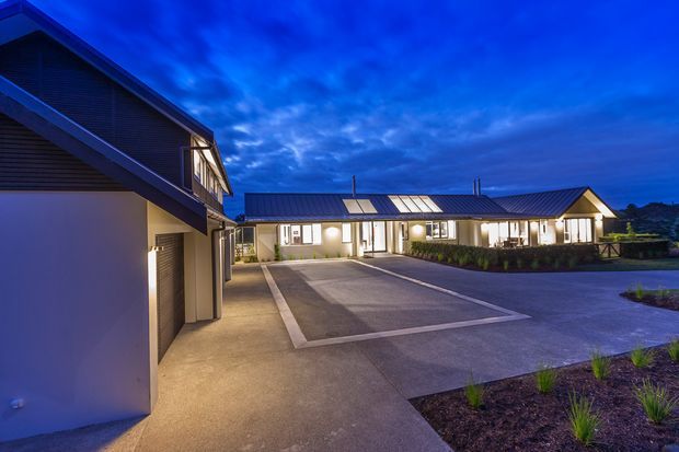 Twilight exterior view of a low-rise contemporary building complex with dark pitched metal roofs, horizontal dark cladding, illuminated windows, covered entries, and a broad paved forecourt with minimal landscaped planting. A larger gabled building occupies the left foreground while a long single-storey wing extends across the background.
