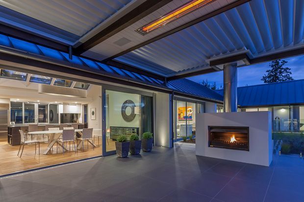 A covered outdoor entertaining area at dusk, with a dramatic linear gas fireplace set into a white feature wall, dark tiled flooring, blue-toned architectural lighting, and a corrugated roof canopy with exposed structural beams. Large sliding glass doors open into a bright dining and kitchen area, while landscaped planting and glazing extend the contemporary indoor-outdoor connection.