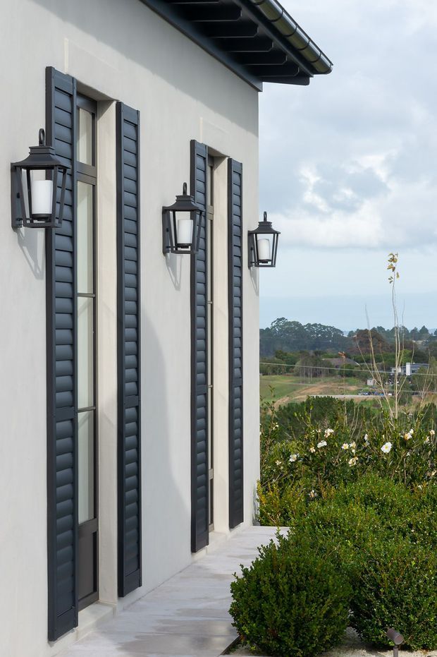 Exterior view of a light-rendered building facade with tall narrow windows, dark louvred shutters, traditional wall-mounted lanterns, exposed guttering, and a pale concrete walkway. Landscaped shrubs and open countryside are visible beside the building.