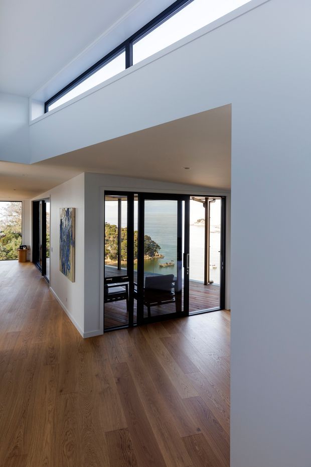A bright, modern interior corridor or living area with warm timber flooring, crisp white walls and ceilings, and extensive black-framed glazing. A dramatic high-level clerestory window brings in natural light above a projecting ceiling plane. Through the glazed doors, a timber deck and a striking coastal water view are visible, with some dark lounge furniture in the adjoining space.