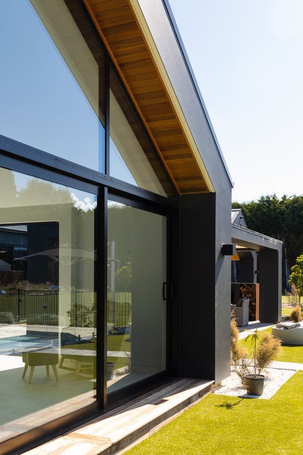 Exterior architectural photograph of a contemporary house extension with expansive black-framed floor-to-ceiling glazing, a strong angular roof overhang with exposed warm timber soffit, dark rendered walls, and a timber threshold opening onto a manicured lawn and gravel garden. A covered outdoor area and parts of the original house are visible in the background.