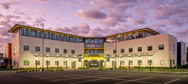 A large contemporary institutional or commercial building photographed from across an empty car park at dusk. The symmetrical two-storey facade is clad in pale masonry, with extensive dark-framed clerestory glazing, projecting angled roof forms, a central recessed entrance with illuminated signage, and linear exterior lighting. Pink and purple evening clouds form the background.