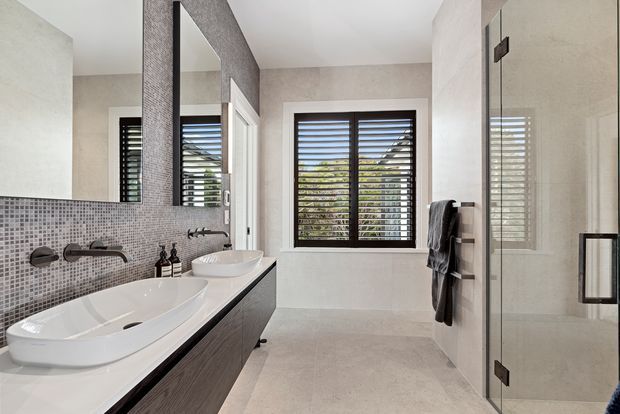 Contemporary bathroom featuring a freestanding white bathtub, wall-mounted dark wood vanity with vessel basin, grey mosaic tile splashback, frameless glass shower enclosure, heated towel rail, and louvred windows. The palette is neutral with textured finishes and clean-lined fixtures.