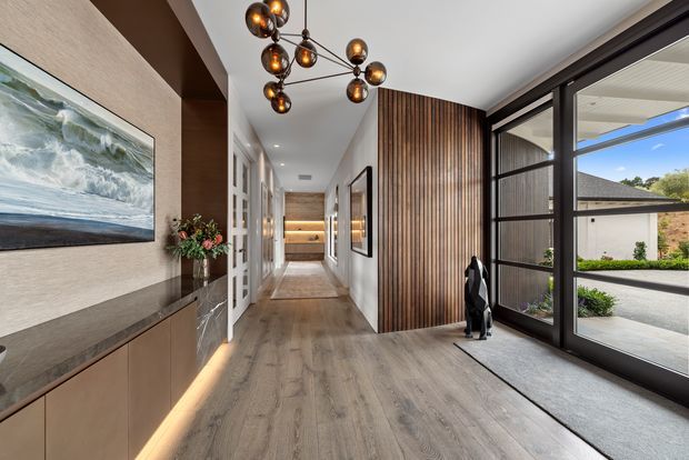 A refined contemporary entry hall with warm timber flooring, a built-in timber and stone console, textured neutral wall finish, large coastal artwork, and a sculptural cluster pendant light. Vertical timber battens define the hallway, while expansive black-framed glazing opens the space visually to the driveway and garden outside. A corridor leads toward additional rooms and illuminated built-in joinery.