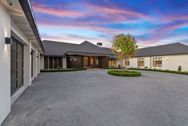 A broad paved courtyard and driveway surrounds a single-storey contemporary residence. The building has dark grey pitched roofs, white rendered walls, dark timber-toned cladding and large windows, with a small landscaped circular garden bed and central tree. The image is taken outdoors at dusk, with warm interior lighting visible through the entry and windows and a colourful sunset sky beyond.