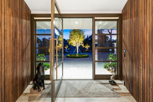 A refined entryway with full-height timber wall panelling, a large glazed pivot door and sidelights opening onto a landscaped courtyard. The space features a neutral stone or concrete floor, a large grey rug, recessed ceiling lighting, and sculptural decorative objects. The courtyard and mature planting are visible beyond the entry.