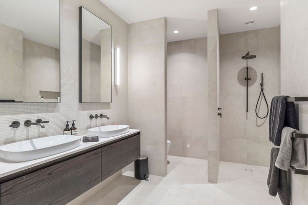 Contemporary double-vanity bathroom with a long dark timber-grain floating cabinet, two white vessel basins, wall-mounted brushed metal taps, large frameless mirrors, and pale stone-look wall and floor finishes. A spacious open shower with a rainfall showerhead and handheld rail is separated by a partial partition wall. Neutral finishes, recessed ceiling lighting, and heated towel rails create a clean, refined atmosphere.