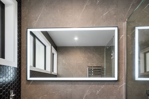Close-up of a contemporary bathroom wall featuring a large rectangular illuminated mirror, brown marble-look wall tiles, a black hexagonal-tile accent strip, black-framed glazing, and a glass shower enclosure reflected in the mirror.