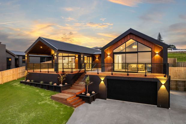 Exterior view of a contemporary single-storey home at dusk, featuring a strong gabled form, dark metal roofing, warm timber-clad facade panels, extensive floor-to-ceiling glazing, and a raised deck with glass balustrades. A large integrated garage sits beneath the main living area, with landscaped lawn, timber fencing, and entry steps in the foreground.