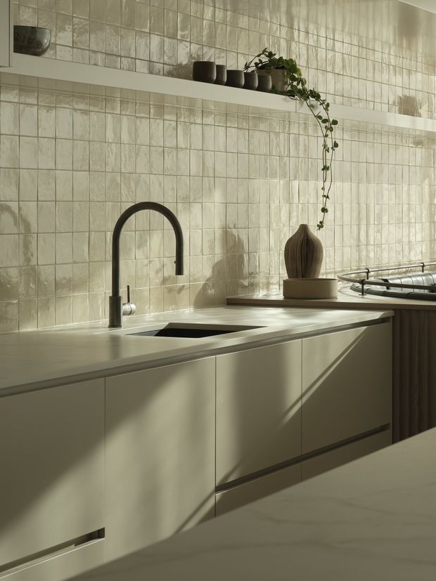 A softly lit contemporary kitchen featuring pale, flush-panel cabinetry, a light stone countertop with an integrated sink, a matte black gooseneck tap, and a textured glossy tile splashback. Open shelving displays dark ceramics and trailing greenery, while warm sunlight creates strong diagonal shadows across the cabinetry and benchtops.