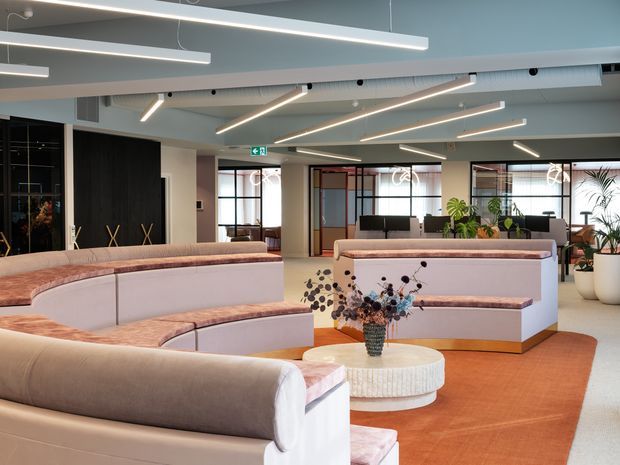 A contemporary commercial lounge or reception area with curved upholstered banquette seating, blush and cream finishes, terracotta carpet, a round textured coffee table, indoor plants, and linear suspended lighting. Glazed meeting or office spaces and dark built-in cabinetry are visible in the background.