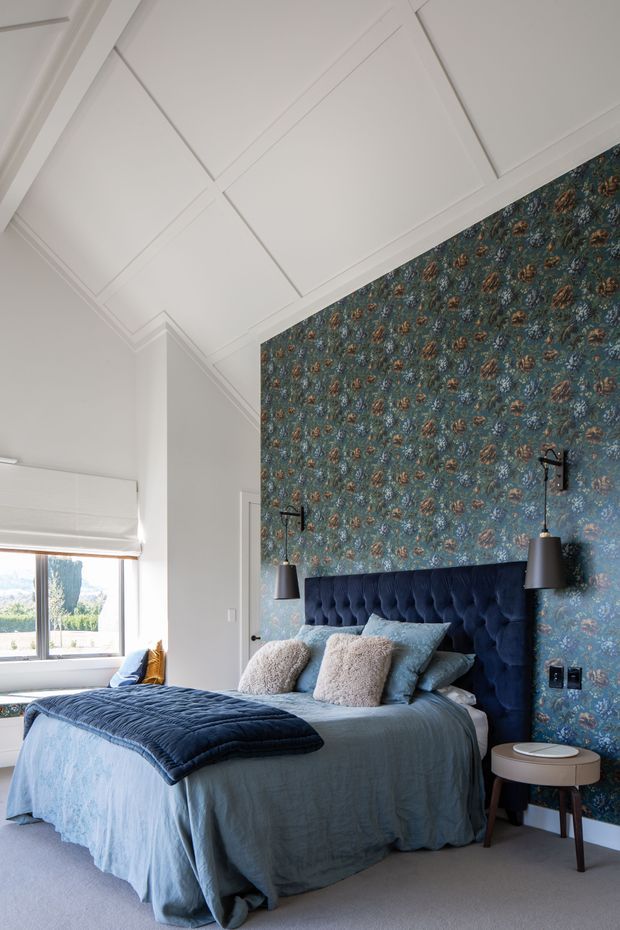 A bright, refined bedroom featuring a deep navy tufted upholstered bedhead, blue-green floral patterned wallpaper, layered blue bedding, textured cream cushions, and matching black wall-mounted pendant lights. White coffered ceiling panels, pale walls, carpeted flooring, and a large window create an airy yet elegant atmosphere.