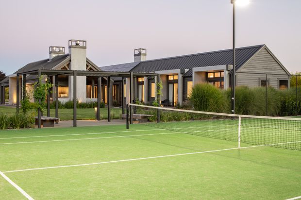 A single-storey residential building with a long, low gabled roof and dark metal roofing is viewed beyond a fenced artificial grass tennis court. The facade combines grey horizontal weatherboard cladding, dark-framed windows and doors, covered outdoor verandah structures, and multiple white rendered chimneys. Landscaping and a lit court occupy the foreground.
