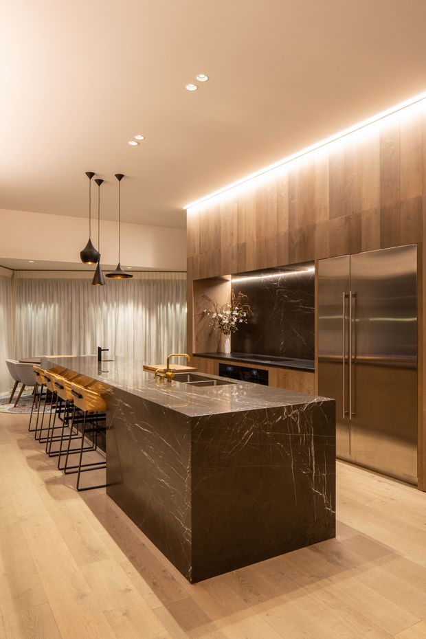 A sophisticated open-plan kitchen and dining area featuring a large dark veined stone island with waterfall edges, integrated cabinetry in warm timber tones, a dark stone splashback, and stainless-steel refrigerator. The island includes a brass tap and seating for several people on slim black-framed stools. Warm concealed LED lighting, pendant lights, pale timber flooring, and full-height sheer curtains create an elegant, softly illuminated interior.