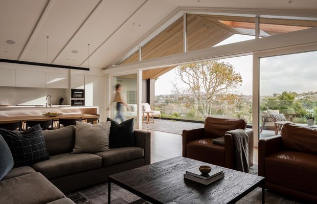 A contemporary open-plan living room with a dark sectional sofa, leather armchairs, and a substantial dark timber coffee table. The space opens directly to a covered outdoor terrace through large sliding glass doors, with a bright white kitchen and island visible behind the seating area. A pitched timber-lined ceiling and extensive glazing create a light, indoor-outdoor connection.