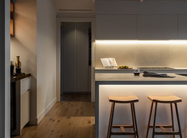 A refined contemporary kitchen viewed from an adjacent hallway, featuring a compact island or breakfast bar with two timber stools, flat-panel grey cabinetry, warm under-cabinet lighting, a pale stone-look benchtop and splashback, and wide-plank timber flooring. A tall concealed storage unit and subtle decorative styling are visible in the background.