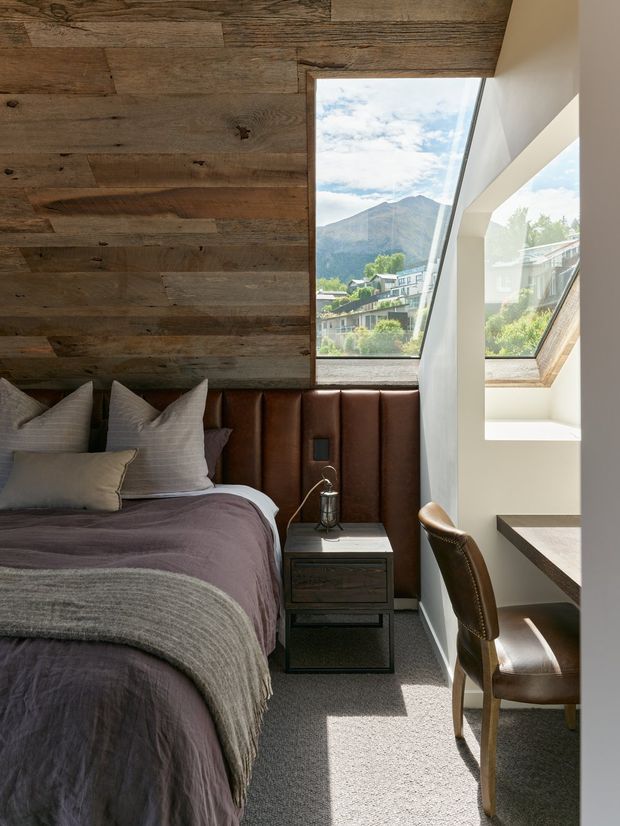 A compact, refined bedroom with a low-pitched timber-lined ceiling and wall, a bed with layered grey and muted purple bedding, and a deep brown upholstered headboard. A narrow bedside table and leather desk chair sit beside a bright built-in workspace. Large angled windows provide views toward a mountainous landscape, while the interior combines warm reclaimed-looking timber, dark upholstery, white plaster and soft natural light.