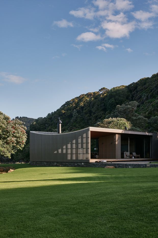A low, modern pavilion-like house set within a broad manicured lawn and wooded hillside landscape. The building has dark vertically ribbed cladding, a shallow flat roof, a stone base, and a deeply recessed covered outdoor living area with large glazed openings. A slender metal chimney rises from the roof, while timber-lined walls and simple outdoor chairs are visible beneath the sheltered verandah.