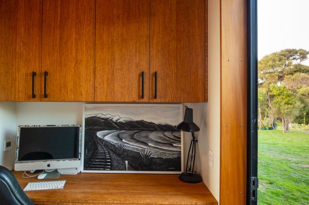 A compact built-in home workspace beside a full-height window or glazed door. Warm timber cabinetry with black handles spans the upper wall, while a matching timber desk supports a computer, keyboard, framed artwork and a black task lamp. The open glazing provides a view to a green garden outside.