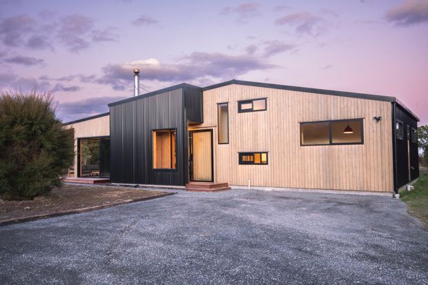 A single-storey contemporary home with a low-pitched roof, clad in pale vertical timber weatherboards and contrasting dark corrugated metal. The facade features asymmetrical projecting volumes, narrow clerestory-style windows, a covered glazed outdoor area, and a gravel driveway and forecourt. The home is photographed at dusk with a pink-purple sky and sparse landscaping.